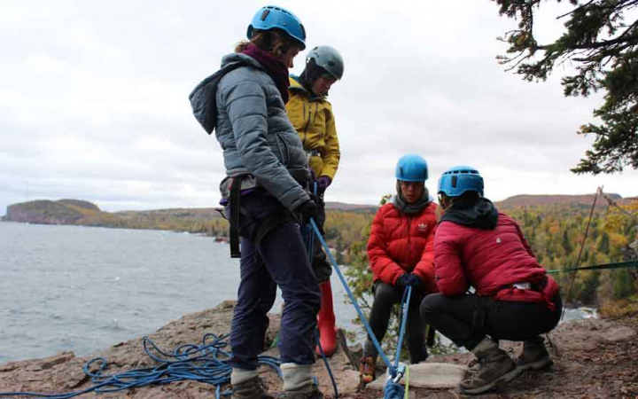 Four people wearing safety gear stand on a cliff above a body of water. Two people are standing, and two are kneeling. 